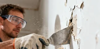 A person using a scraper to remove old paint from walls while wearing safety gloves and goggles during home renovation.