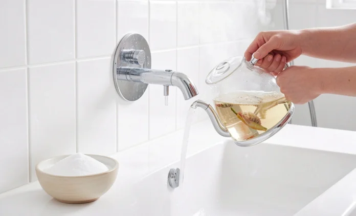 Person using boiling water and baking soda to unclog shower drain naturally in bright white bathroom Person using boiling water and baking soda to unclog shower drain naturally in bright white bathroom