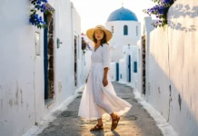 20 Best Greece Trip Outfits: Your Santorini to Mykonos Style Woman in white linen dress and straw hat on Santorini cobblestone street — greece trip outfits