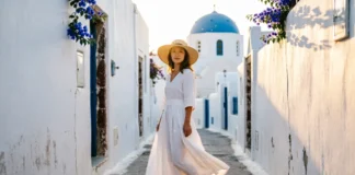 Woman in white linen dress and straw hat on Santorini cobblestone street — greece trip outfits