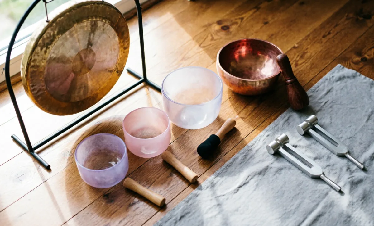 Collection of sound healing instruments including a gong, crystal singing bowls, Tibetan bowl, and tuning forks on a wooden floor.
